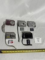 Lot of five digital cameras including Nikon, Sony, and Canon models with some batteries shown. Cameras arranged with measuring tape.