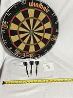 Front view of Winmau Blade 4 dartboard with 3 darts placed below, along with measuring tape for size reference.
