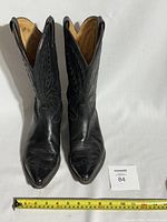 Front view of the pair of black leather cowboy boots showing pointed toes, detailed stitching on the shaft and vamp, and measuring tape for size.