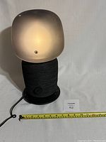 Full view of the Sonos IKEA SYMFONISK table lamp speaker lit from inside with a frosted glass shade, black woven fabric base, power cord visible, measuring tape showing height.