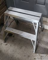 Full view of aluminum ladder showing two wide steps and A-frame legs, placed indoors on concrete floor.