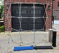 Photo showing one foldable metal display frame fully expanded outside, with blue and black carry bags and additional collapsed frames nearby.