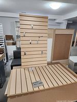 Two brand new light wood melamine slatwall panels with horizontal slat grooves. Some metal hooks attached to one panel, panels shown leaned up and laid flat.