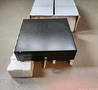 Top view of black Vesario cash drawer with key in lock, resting on white foam supports and on a floor with original box behind it.