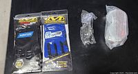 Photo showing two unopened pairs of Mechanix Wear gloves (one black Norton Medium, one blue and black Speed Grip X-Large), one pair of clear safety glasses in plastic packaging, and one pair of clear safety goggles in plastic packaging.