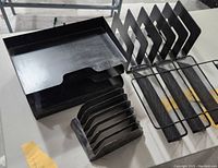 Two black plastic two-tier desk trays, black plastic letter sorter with eight slots, and three black metal mesh vertical file organizers on a glass table.