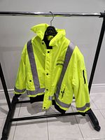 Full front view of the bright yellow winter security jacket hanging on a black metal rack, showing reflective silver stripes on front and sleeves, with black pockets and cuffs.