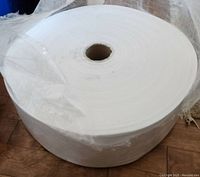 Large roll of white Anmolfabtex fabric wrapped in clear plastic showing fabric wound around a cardboard core.