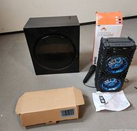 Lot contents: Samsung subwoofer, black portable wireless speaker with blue LEDs, wired handheld microphone, instruction pamphlet, and plain cardboard box likely containing the battery