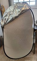 Oval photography reflector with white diffusion panel and silver reflective cover partially draped over top edge, standing upright indoors near window.