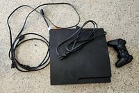 Top view of PS3 Slim console with power and HDMI cords and one DualShock 3 controller on a light floor surface.