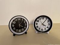 Two small round clocks side by side, one with black face and silver numbers, the other with white face and black numbers, metal rim and two small feet