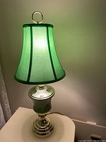 Front view of the brass lamp with green ceramic base and green fabric shade lit up, showing the lamp on a white surface against a pale wall.