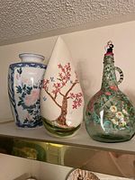 Three vases side by side on a shelf, showing the entire group including the floral and bird designs on the ceramics and the green glass bottle shape with handle and stopper