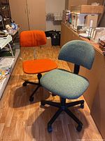 Photo showing both green and orange desk chairs side by side on a wooden floor in a cluttered storage/office space.