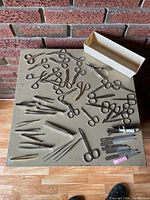 Overview of the lot showing various eye tweezers, small nail scissors, nail files, and a white storage box placed on a table against a brick wall.