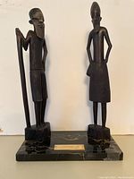 Front view of two hand carved wooden bearded men sculptures mounted on a black marble base with a label indicating made in Kenya.