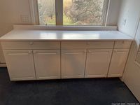 Photo showing front view of three white cupboard units under a white countertop with a backsplash edge against a wall with window.