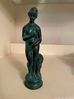 Front and 3-quarter view of green metal figurine of a woman in classical Greek style with detailed base.