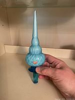 Front view of the ornate blue glass wine stopper held in hand, showing the textured bulbous center, tiered pointed finial, and cylindrical base.