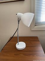 Photo of the white plastic desk lamp on a wood surface next to a window with blinds, showing the lamp shade and part of the flexible neck.