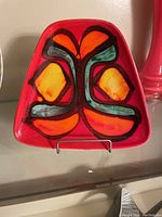 Front view of a red triangular ceramic plate with abstract orange, yellow, and teal pattern outlined in black.