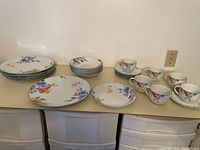Photo showing the full set arranged in groups on a table, displaying plates, bowls, cups, and saucers with floral designs.