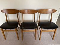 Front view of three teak wood dining chairs with black vinyl seats, one seat shows signs of damage.