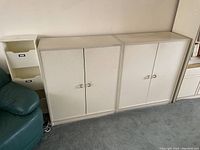 Two white medium-sized storage cabinets positioned side by side on a carpeted floor next to a white shelving unit and green armchair.