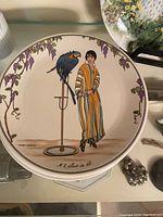 Plate with woman wearing striped orange and yellow robe and a parrot on a stand, grapevine border