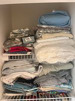 Closet shelf stacked with a variety of folded linens including quilts, blankets, sheets, and pillowcases in neutral colors and patterns.
