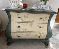 Front view of bow-front chest of drawers showing floral vine painted drawer fronts and decorative top edges.