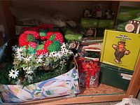 Photo showing holiday garland with red strawberry accents, white flowers, and a box of string lights stacked with other holiday decorations.