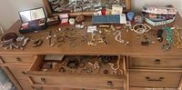 Wide view of dresser top displaying various costume jewelry items including bracelets, necklaces, pins, and coins in cases across the surface.