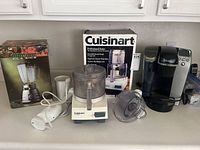 Photo showing all items lined up on a white countertop: boxed Hamilton Beach blender, white hand mixer, Cuisinart food processor next to its box, Keurig coffee maker.