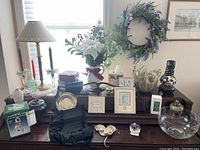 Photo of top of piano showing porcelain figurines, candlesticks, vases, picture frames, and other decorative items with lamp, floral arrangement, and wreath in background.