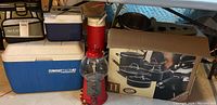 Photo showing a red gumball machine, blue Coleman cooler, white cooler, and a boxed 11-piece cookware set underneath a table.