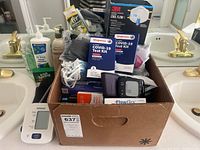 Photo showing cardboard box filled with health and toiletry items including COVID-19 test kits, blood pressure monitors, and masks