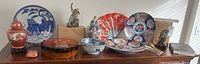 Wide shot of Japanese antiques and collectibles displayed on top of a wooden cabinet including plates, fan, figurine, jar, and box.