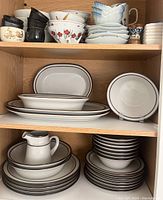 Full view of open shelf showing multiple pieces of Japanese stoneware dinnerware, stacked plates, bowls, small cups with floral patterns, and blue geometric designs.