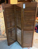 Three-panel wooden room divider with louvered slats shown partially folded in a basement setting with boxes and other storage items visible.