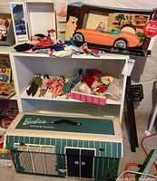 Full view of white shelves with Barbie dolls, clothing, car in original box, and Dream House carrying case on floor below