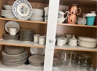 Wide view of multiple shelves showing stacked plates, bowls, cups, creamers, sugar set, and glassware including clear glasses and copper mugs