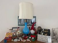 Overview of table top showing blue glass lamp with beige shade, assorted small items, fabric gnomes, and decorative container with a face.