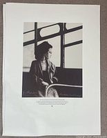 Photo showing monochrome print of a woman seated near a window, signed by the artist, with text beneath the artwork.