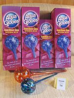 Four Aqua Globes watering bulbs, three large in boxes and one smaller bulb loose, displayed on couch