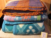 Photo showing three folded blankets stacked. Bottom is a large blue fleece blanket with bold turquoise and white patterns. Middle is a medium blue fleece with floral or circular pattern. Top is coarse wool blanket in purple and orange plaid with orange blanket-stitch edge.