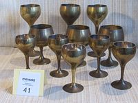 Photo showing the full set of 11 brass goblets arranged in three rows according to size with five small in front, three medium in middle, and three tall in back. The goblets show tarnishing and signs of usage.