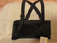 Black Kuny elastic support belt with wide elastic bands and velcro closure, featuring two adjustable elastic suspenders crossing at back, laid on beige couch.