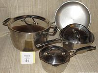 Grouped photo showing three stainless steel pots and one frying pan placed on a fabric surface. One lid is mismatched and made of glass.